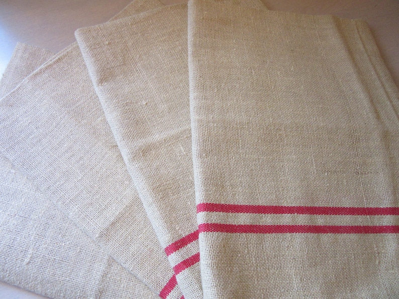 May include: Four natural linen tea towels, one with two red stripes. The towels are folded and stacked on top of each other.