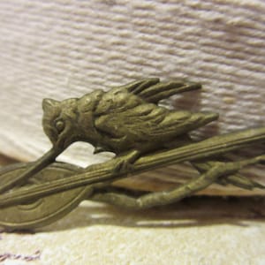 May include: A vintage-style brass brooch featuring a bird perched on a stringed instrument. The bird has a long beak and detailed wings. The brooch is a warm, antique gold color, with intricate detailing on both the bird and the instrument.
