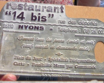 May include: A silver metal sign with the text "restaurant "14 bis" 26110 NYONS rue des déportés Téléphone : (75) 26, 00.45 Ouvert tous les jours (midi et soir) (sauf mardi et dimanche midi) Plats du Jour - Spécialités Réugoundines Carte - Possibilité Menu par Commande Registre du Commerce en cours N° d'identification NYONS"