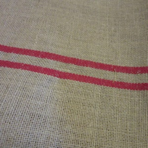 May include: A natural burlap fabric with two red stripes running horizontally across the fabric.