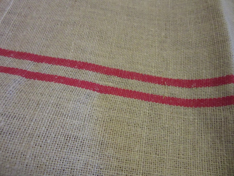 May include: A natural burlap fabric with two red stripes running horizontally across the fabric.