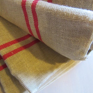 May include: Two pieces of beige burlap fabric with red stripes folded on top of each other. The fabric has a rough texture.