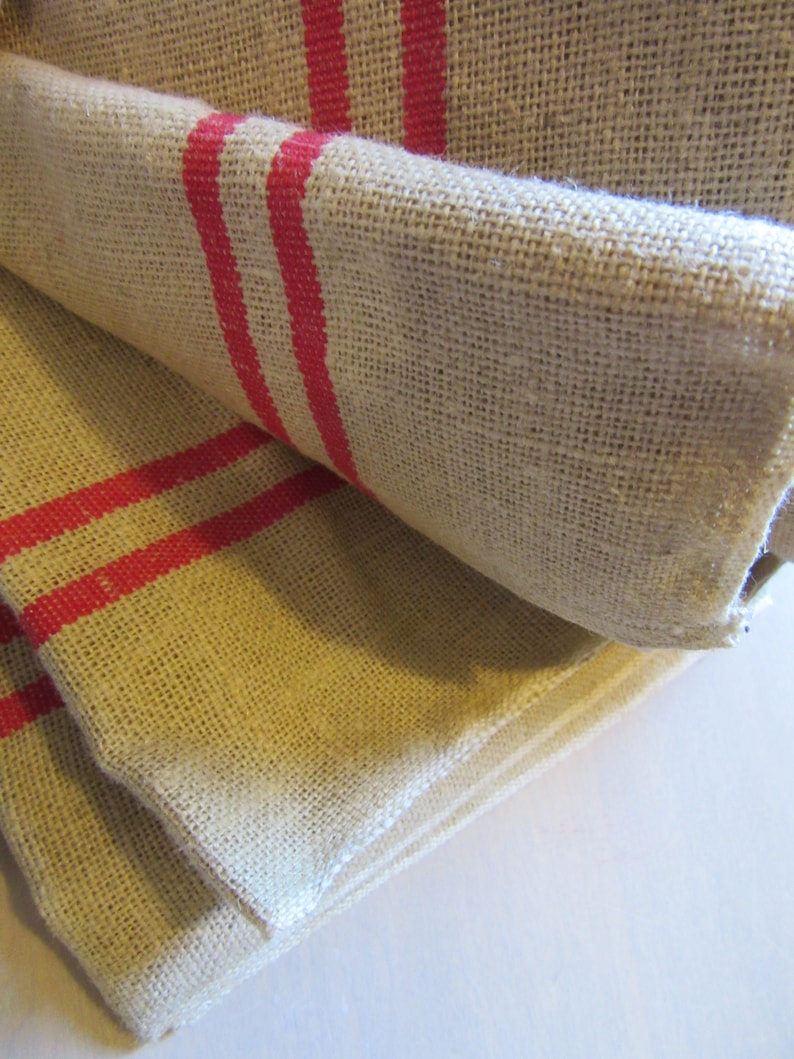 May include: Two pieces of beige burlap fabric with red stripes folded on top of each other. The fabric has a rough texture.