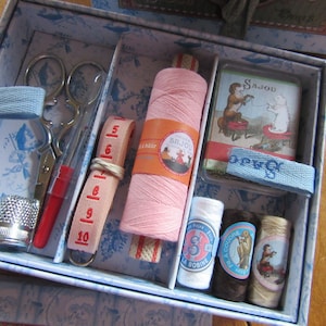 May include: A vintage sewing kit in a blue floral patterned box. The kit includes scissors, a thimble, measuring tape, pink thread, and spools of thread. A small card with a vintage illustration is also included.