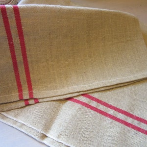 May include: Two natural linen tea towels with red stripes. The towels are folded and stacked on top of each other.