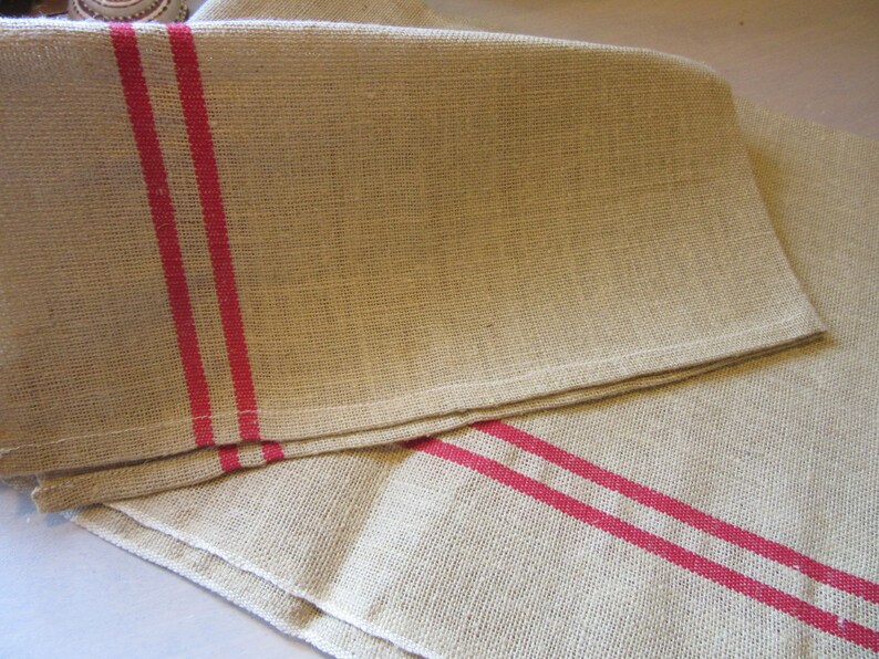 May include: Two natural linen tea towels with red stripes. The towels are folded and stacked on top of each other.