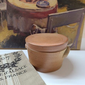 May include: A round, tan-colored ceramic crock with a matching lid and two small side handles. Next to the crock is a beige fabric cloth featuring a black printed design with the text "de" and "D'ALSACE S, FRANCE" visible on the fabric surface.