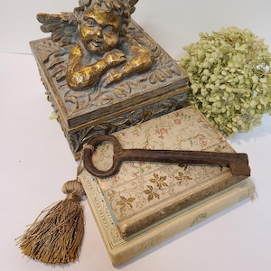 Puede incluir: Arreglo decorativo de estilo antiguo. Incluye una caja de querubín con detalles dorados, una llave grande y oxidada y una pila de libros antiguos. Una borla dorada y flores secas completan la composición. La caja mide unos 15 cm de largo.