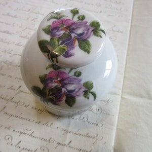 May include: White porcelain jar with a floral lid. The lid features a purple and white flower design. The jar has a similar floral design with purple and white flowers.