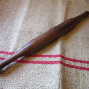 May include: A wooden rolling pin with a smooth, dark brown finish. The rolling pin is lying on a beige linen fabric with red stripes.