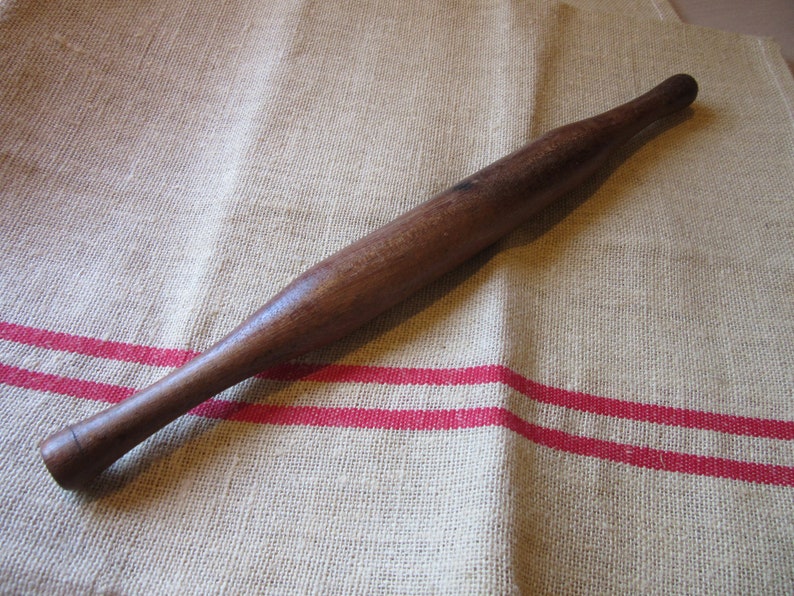 May include: A wooden rolling pin with a smooth, dark brown finish. The rolling pin is lying on a beige linen fabric with red stripes.