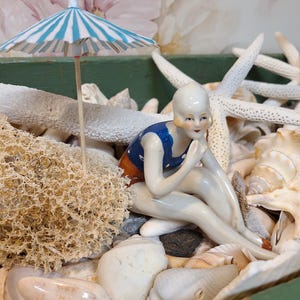 Könnte beinhalten: Vintage Porzellanfigur einer Frau in blauem Badeanzug, die zwischen Muscheln, Seesternen und einem Miniatur-Streifenschirm sitzt. Die Figur hat eine klassische, retro Ästhetik und erinnert an ein Strandthema.