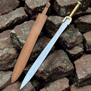 Op de afbeelding: Een Romeins gladius-zwaard met een gepolijst stalen lemmet en een messing gevest. Het zwaard wordt getoond met zijn bruine leren schede, rustend op een bed van ruwe, roodbruine stenen. De schede heeft decoratieve stiksels.