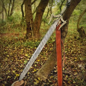 May include: A long, ornate sword with a patterned blade and decorative hilt, resting against a tree. The sword has a brown leather sheath. The background features a forest setting with trees and fallen leaves.
