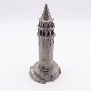 Raw finish Galata Tower miniature in unpolished cast stainless steel seen from a slight angle, showing the round base and conical roof on a white background