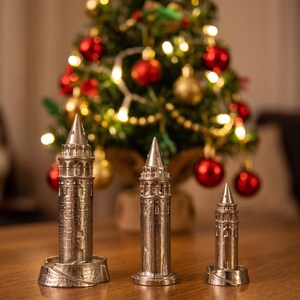 Three polished Galata Tower miniatures in small, medium and large sizes on a wooden table in front of a lit Christmas tree