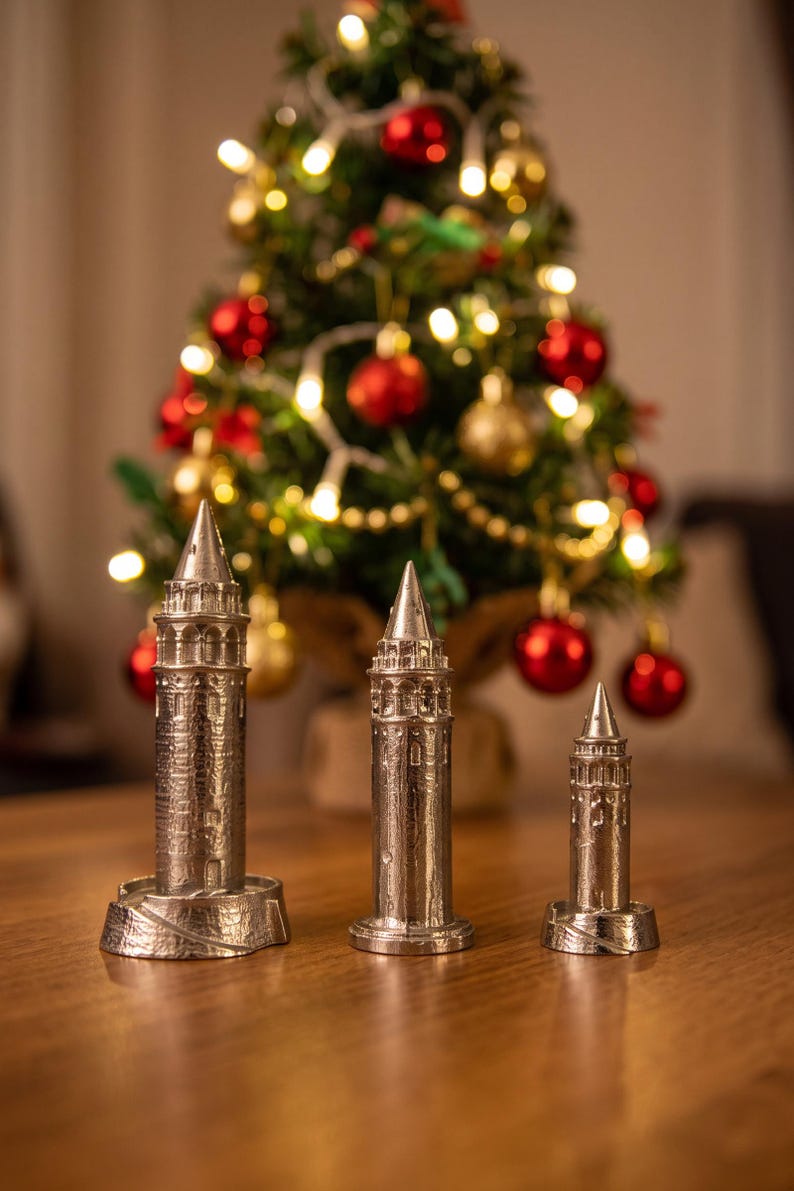 Three polished Galata Tower miniatures in small, medium and large sizes on a wooden table in front of a lit Christmas tree