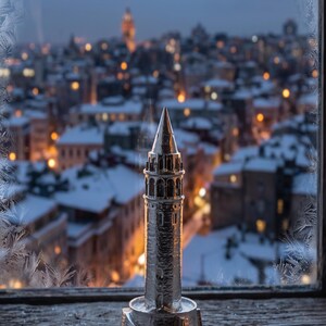 Polished stainless steel Galata Tower miniature on a window sill with frost on the glass and a snowy Istanbul skyline at dusk behind it