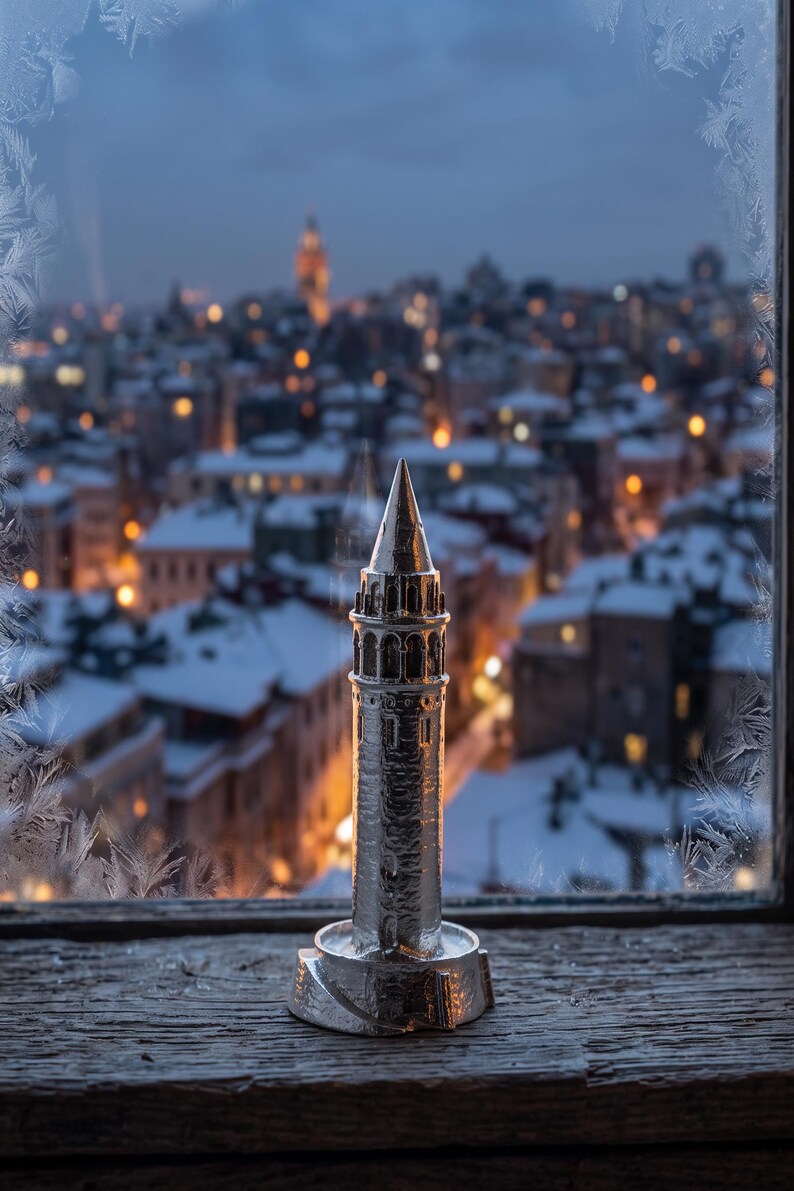 Polished stainless steel Galata Tower miniature on a window sill with frost on the glass and a snowy Istanbul skyline at dusk behind it