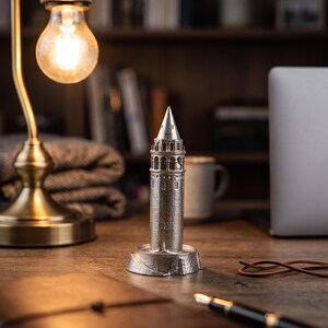Galata Tower miniature on a desk beside a leather notebook and a fountain pen under a glowing bulb lamp