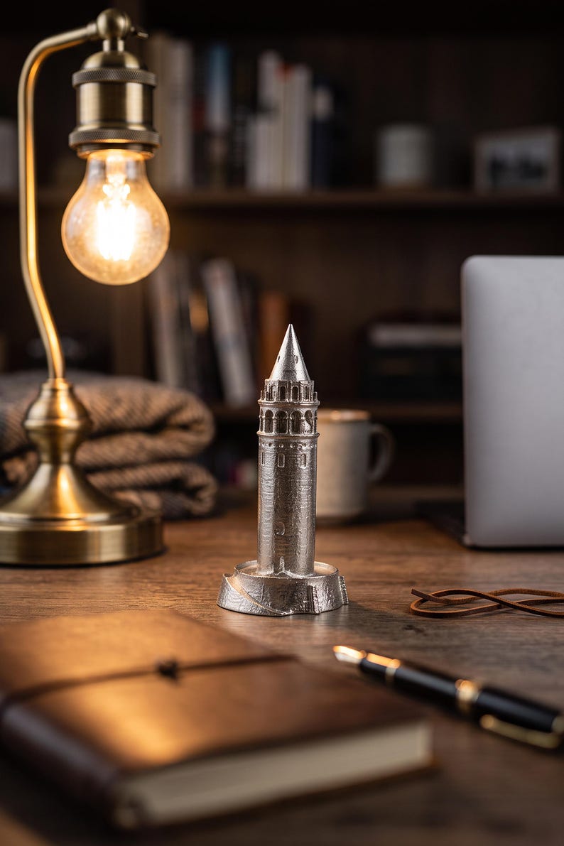 Galata Tower miniature on a desk beside a leather notebook and a fountain pen under a glowing bulb lamp