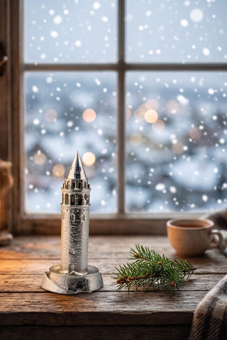 Polished Galata Tower miniature on a rustic window sill with snow falling outside, a cup of tea and a pine branch