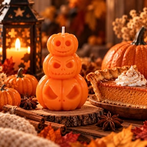 May include: A stack of three orange pumpkin-shaped candles with carved facial features, positioned on a wooden slice. The candles feature a smooth wax texture and a single white wick at the top, surrounded by autumn-themed decor.