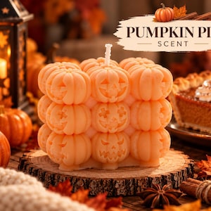 May include: A square-shaped orange candle composed of nine individual pumpkin-shaped segments, each featuring a unique carved jack-o'-lantern face. The candle has a single white wick at the top and is labeled with text reading PUMPKIN PIE SCENT.