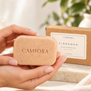 May include: A rectangular bar of light brown cinnamon soap with the word CAMFORA embossed on the front. Beside it sits a brown cardboard box with a white label reading CAMFORA CINNAMON NATURAL HANDMADE SOAP 3.5 oz (100 g).