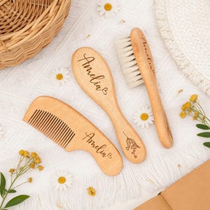 Könnte beinhalten: Ein Set aus drei Holz-Haarpflegeartikeln: ein Kamm, eine ovale Bürste und eine Bürste mit Borsten. Jeder Artikel ist mit dem Namen "Amelia" graviert. Die Artikel sind auf einer weißen Oberfläche mit Gänseblümchen dekoriert, für Babys.