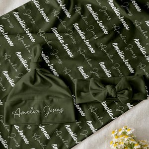 May include: Olive green baby blanket, hat, and headband set. The blanket and hat are printed with the name "Amelia Jones" in a repeating pattern. The headband features a bow. The set is designed for newborns.