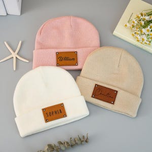 Könnte beinhalten: Drei personalisierte Mützen in Rosa, Creme und Beige. Jede Mütze hat einen braunen Lederaufnäher mit einem Namen: "William", "Sophia" und "Christian". Die Mützen sind am Rand gefaltet.