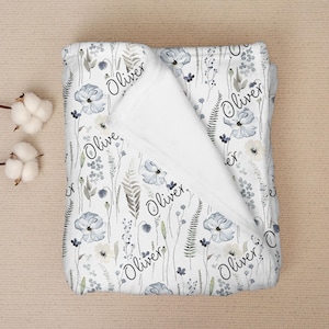 Könnte beinhalten: Eine weiße Babydecke mit einem floralen Muster in Blau-, Grau- und Cremetönen. Die Decke trägt den Namen "Oliver" in einer Schreibschrift, der sich über das Design wiederholt. Die Decke ist gefaltet und zeigt ein weiches, weißes Innenfutter.