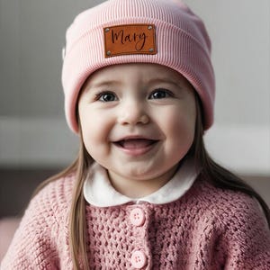 May include: A light pink knit beanie with a brown leather tag that reads "Mary" in cursive. The beanie is worn by a smiling child, who is also wearing a pink knit cardigan with a white collar and pink buttons.