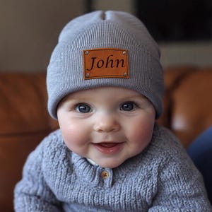 Könnte beinhalten: Graue Strickmütze mit braunem Lederaufnäher mit dem Namen "John". Das Baby trägt einen grauen Pullover. Das Baby hat blaue Augen und lächelt. Ein beliebtes Babyaccessoire.