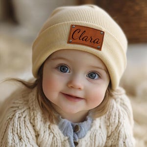 Könnte beinhalten: Hellbeige Strickmütze mit einem braunen Lederaufnäher mit dem Namen "Clara" in Schreibschrift. Die Mütze wird von einem Baby getragen, das einen cremefarbenen Pullover trägt.