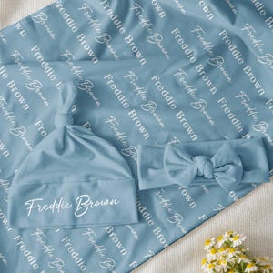 Könnte beinhalten: Ein hellblaues Baby-Set mit Decke, Mütze und Stirnband. Der Name "Freddie Brown" ist in weißer Schrift auf Decke und Mütze gedruckt. Das Stirnband hat eine Schleife. Das Set wird auf einer weißen Oberfläche präsentiert.