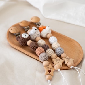 Puede incluir: Pinzas de chupete de madera para bebés con cabezas de animales de ganchillo, incluyendo un oso y un zorro. Las pinzas tienen cuentas de ganchillo en colores neutros y colgantes de estrella de madera grabados con nombres. Se muestran en una bandeja de madera.