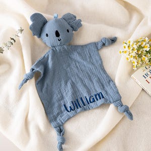 Könnte beinhalten: Eine blaue Koala-förmige Baby-Kuscheldecke mit dem Namen "William" auf der Vorderseite gestickt. Die Decke hat geknotete Arme und Beine und eine kleine Schlaufe oben. Der Koala hat schwarze Augen und einen lächelnden Mund.