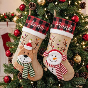 Könnte beinhalten: Zwei beige Weihnachtsstrümpfe aus Jute mit rot-schwarz karierten Bündchen. Ein Strumpf zeigt ein Plüsch-Rentier mit grün gestreiftem Schal und dem Namen Anna. Der andere zeigt einen Schneemann mit rot gestreiftem Schal und dem Namen Frank.