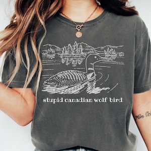 Pode incluir: Camiseta cinza escuro com um gráfico branco de um mergulhão em um lago com árvores e montanhas. O texto "stupid canadian wolf bird" está impresso abaixo. A camiseta tem mangas curtas.