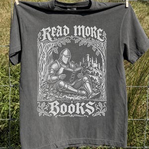 May include: A charcoal gray short-sleeve t-shirt featuring a white graphic print of a knight in armor sitting under a tree reading a book with a castle in the background. The text READ MORE is printed above the knight and BOOKS is printed below.