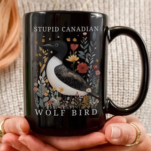 May include: Black ceramic mug with a large handle, featuring a detailed illustration of a loon bird amidst colourful flowers and foliage. The mug has the text "STUPID CANADIAN" above the bird and "WOLF BIRD" below.