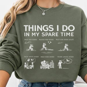 May include: Olive green sweatshirt with white text that reads "THINGS I DO IN MY SPARE TIME." The graphic includes illustrations of bird watching activities. A casual, comfortable top for bird enthusiasts.