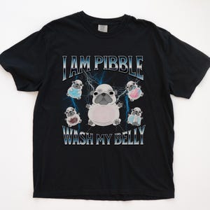 Pode incluir: Camiseta preta com um gráfico apresentando um pug de desenho animado e o texto "I AM PIBBLE WASH MY BELLY". O design inclui raios e vários pugs menores vestindo roupas.