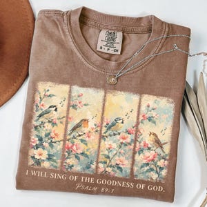 Puede incluir: Una camiseta marrón de manga corta con un gráfico de cuatro paneles verticales con pájaros sobre ramas florecidas. El texto "I WILL SING OF THE GOODNESS OF GOD. Psalm 89:1" está impreso debajo de los paneles. Un collar plateado descansa sobre la prenda.