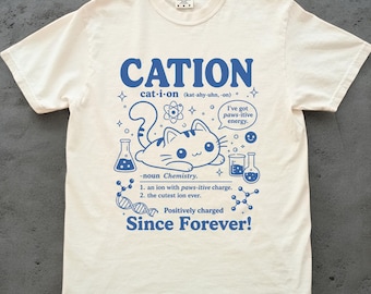 May include: A cream-colored short-sleeve t-shirt featuring a blue graphic print. The design includes a cartoon cat, chemistry equipment, and text that reads CATION, cat-i-on, I've got paws-itive energy, noun Chemistry, an ion with paws-itive charge, the cutest ion ever, Positively charged Since Forever!