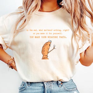 May include: Cream-colored t-shirt with the text "In the end, what matters? nothing, right? so you make it for yourself. YOU MAKE YOUR RIGATONI PASTA." A cartoon pasta character is also featured.