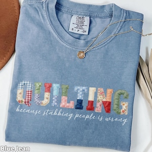 Op de afbeelding: Blauw spijkerbroekkleurig T-shirt met het woord "QUILTING" in kleurrijke patchwork letters. De tekst "because stabbing people is wrong" staat eronder. Een gouden ketting met hanger ligt op het shirt.