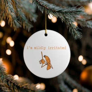 May include: A white ceramic Christmas ornament with the text "i'm mildly irritated" in a brown font. The ornament features a cartoon illustration of a brown insect holding a stick. A gold string is attached for hanging.